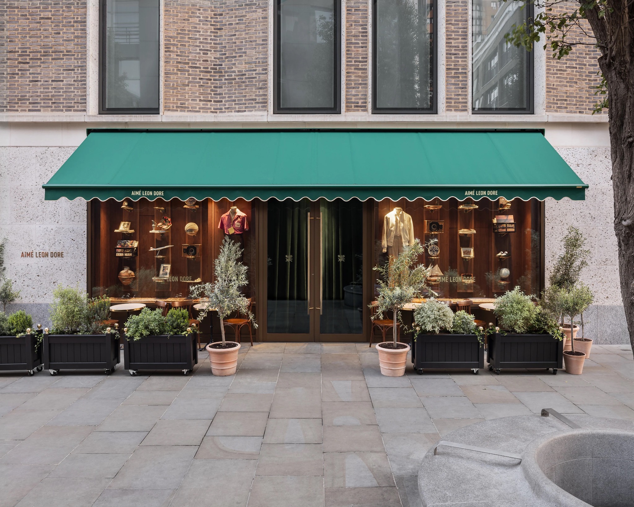 The exterior of the Aimé Leon Dore London flagship with green awning, café seating and planting